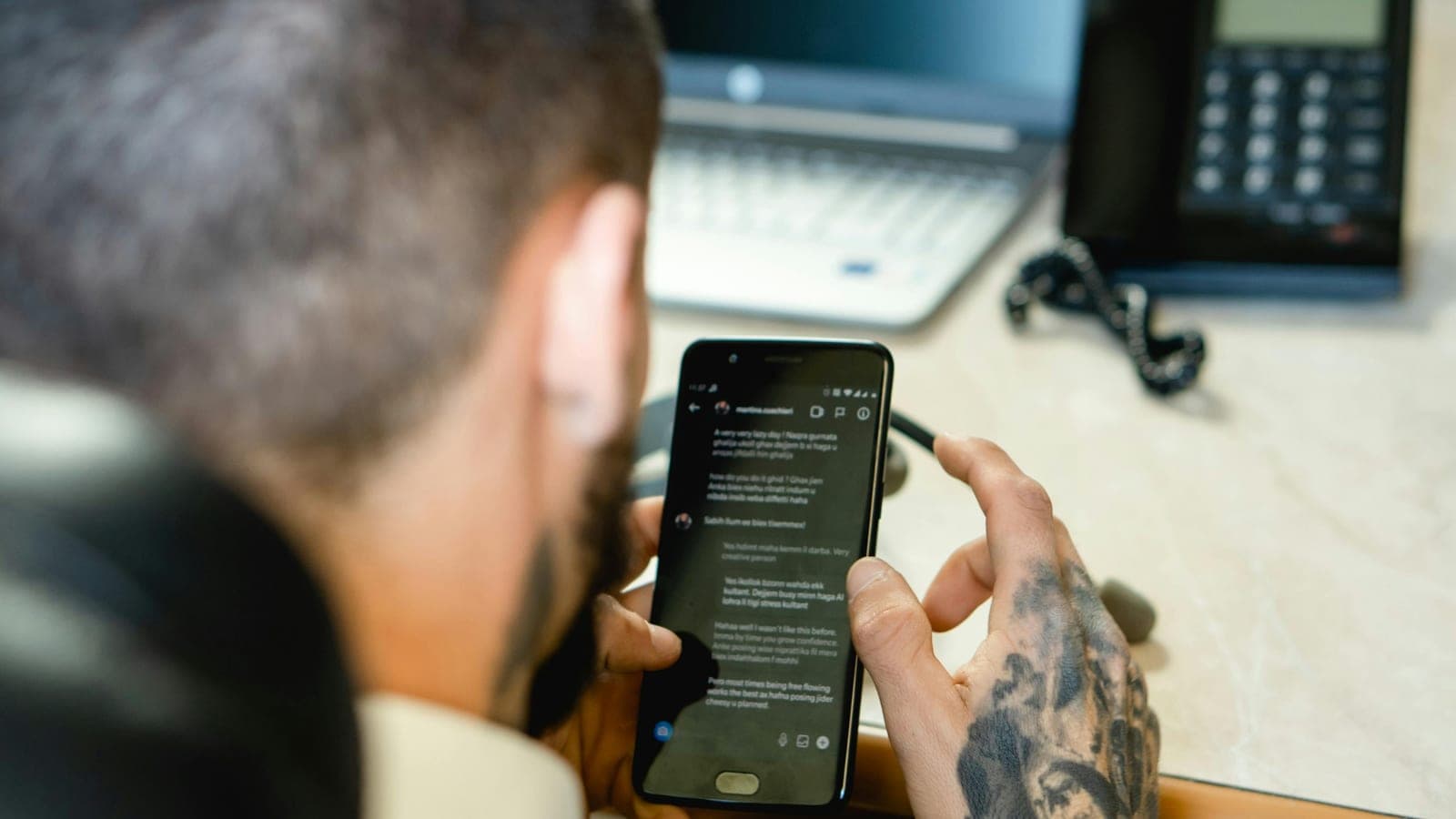Tatuador sentado a uma secretária a consultar mensagens no telemóvel com um portátil aberto ao lado