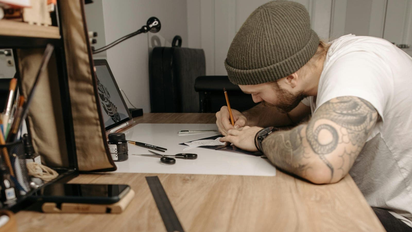 Tattooed forearms at a wooden studio desk with a stencil and a ruler