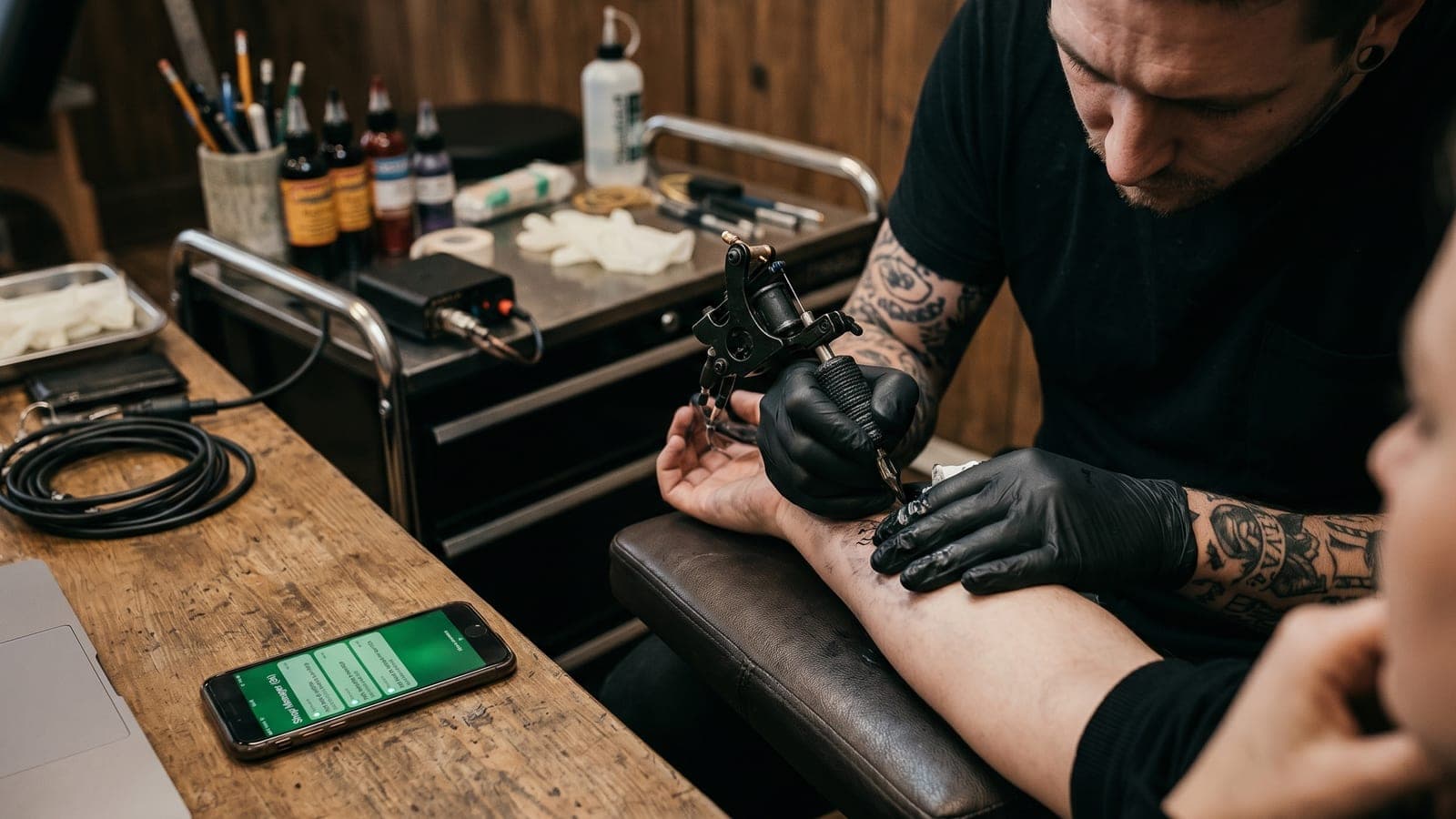 A tattoo artist at work with a phone on the bench showing a client conversation